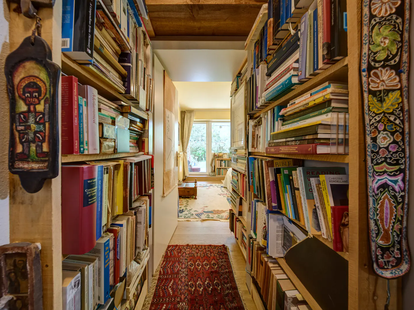 Hallway lined with floor-to-ceiling bookshelves filled with books. A red rug runs down the center, leading to a bright room with a window.