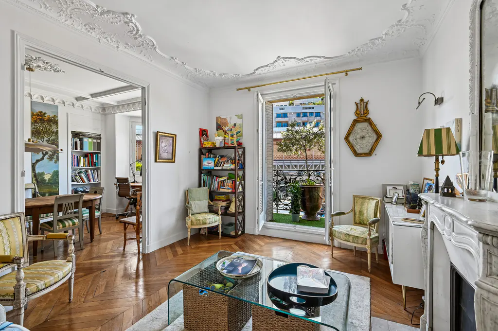 Bright living room with herringbone wood floors, white walls, and ornate ceiling molding. Balcony with potted tree visible through open doors.