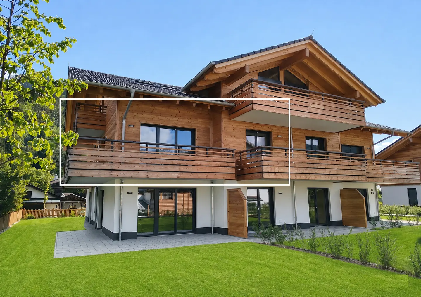 Two-story house with wood siding, black windows, and balconies on both floors. Green lawn and blue sky.