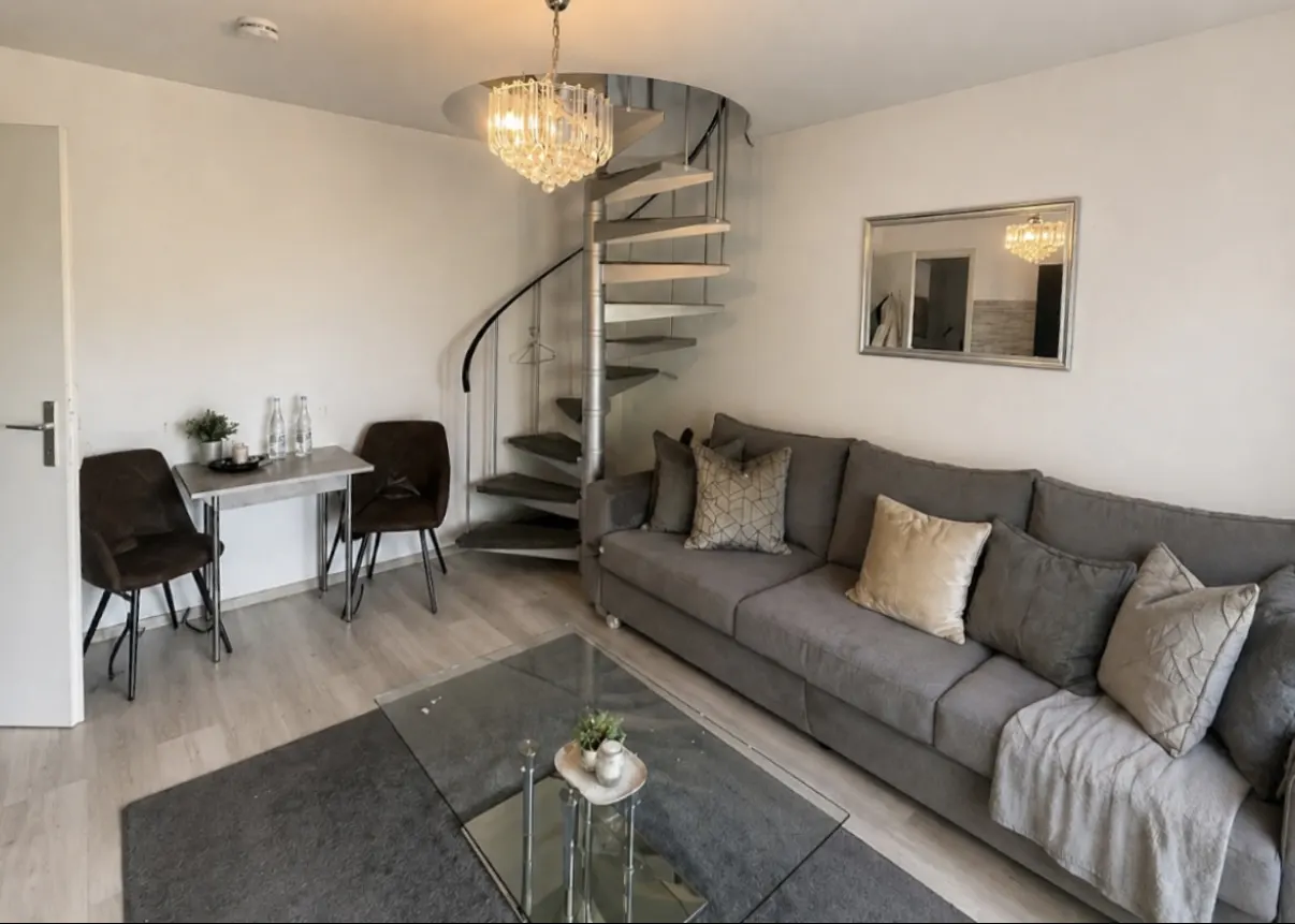 Living room with gray sofa, glass table, and spiral staircase. Chandelier hangs above the stairs. A mirror reflects the room.