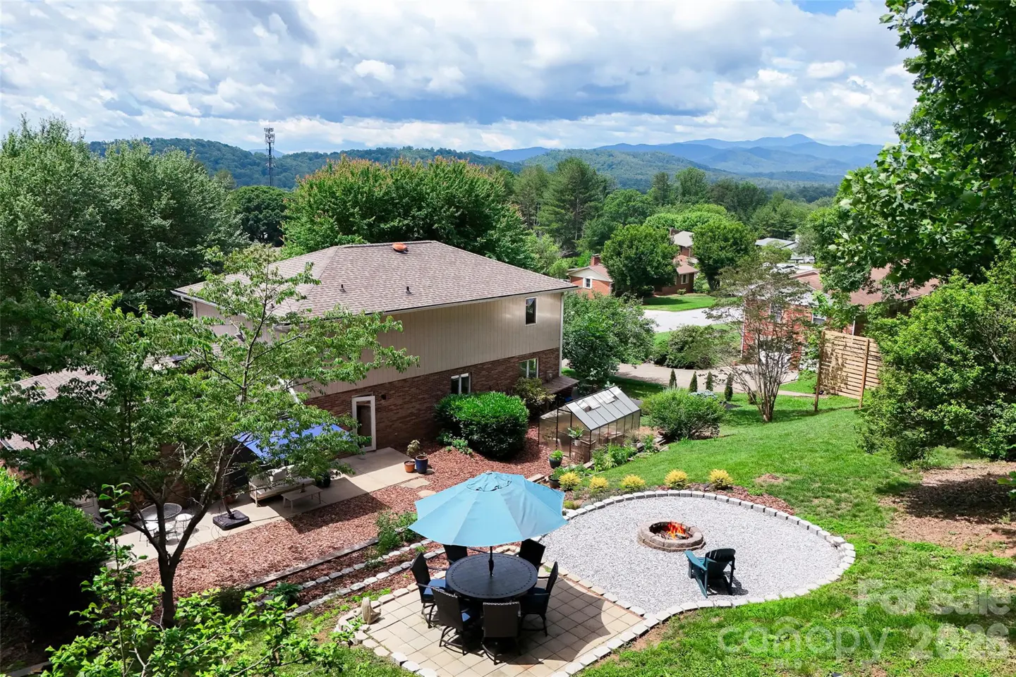Backyard view of a house with a patio, blue umbrella, dining set, fire pit, and a greenhouse overlooking mountain views.