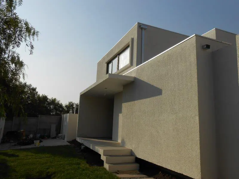 Modern, two-story house with a textured, light-gray exterior. Steps lead to a covered entrance. Green lawn and trees in the background.