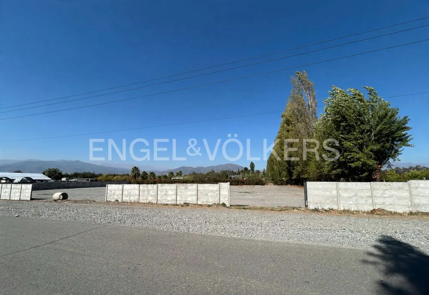 A vacant lot is enclosed by a gray concrete wall, with trees and mountains in the background under a blue sky.
