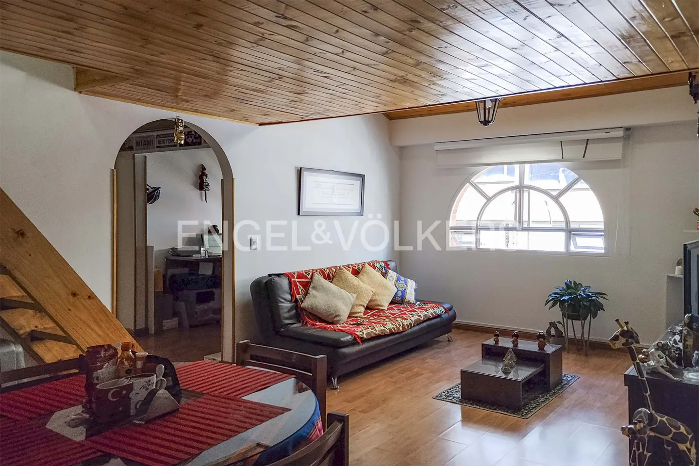 Living room with wood ceiling, black sofa with patterned throw, coffee table, and arched window. Dining table in foreground.