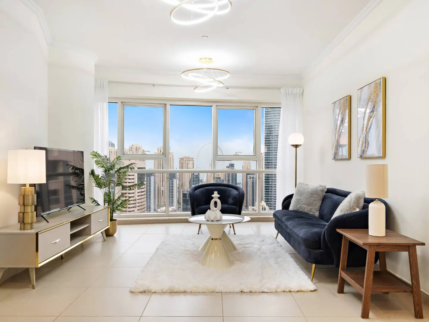 Bright living room with a blue sofa, chair, and a large window overlooking a city skyline. A white rug and modern lighting complete the space.