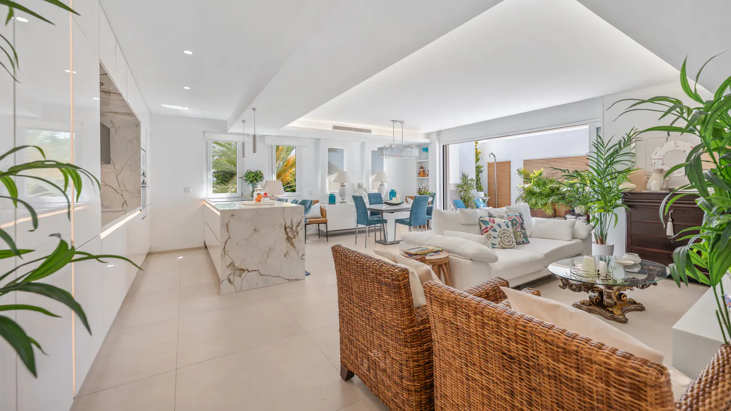 Modern open-plan living area with white marble kitchen island, white sofa, wicker armchairs, and indoor plants.