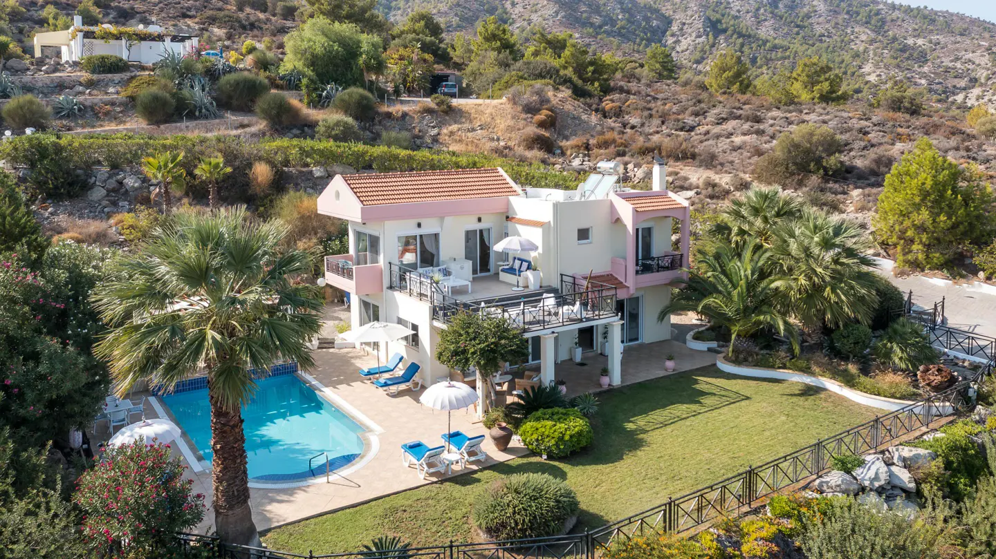 Aerial view of a two-story house with a pink roof, a pool, palm trees, and a mountain backdrop.