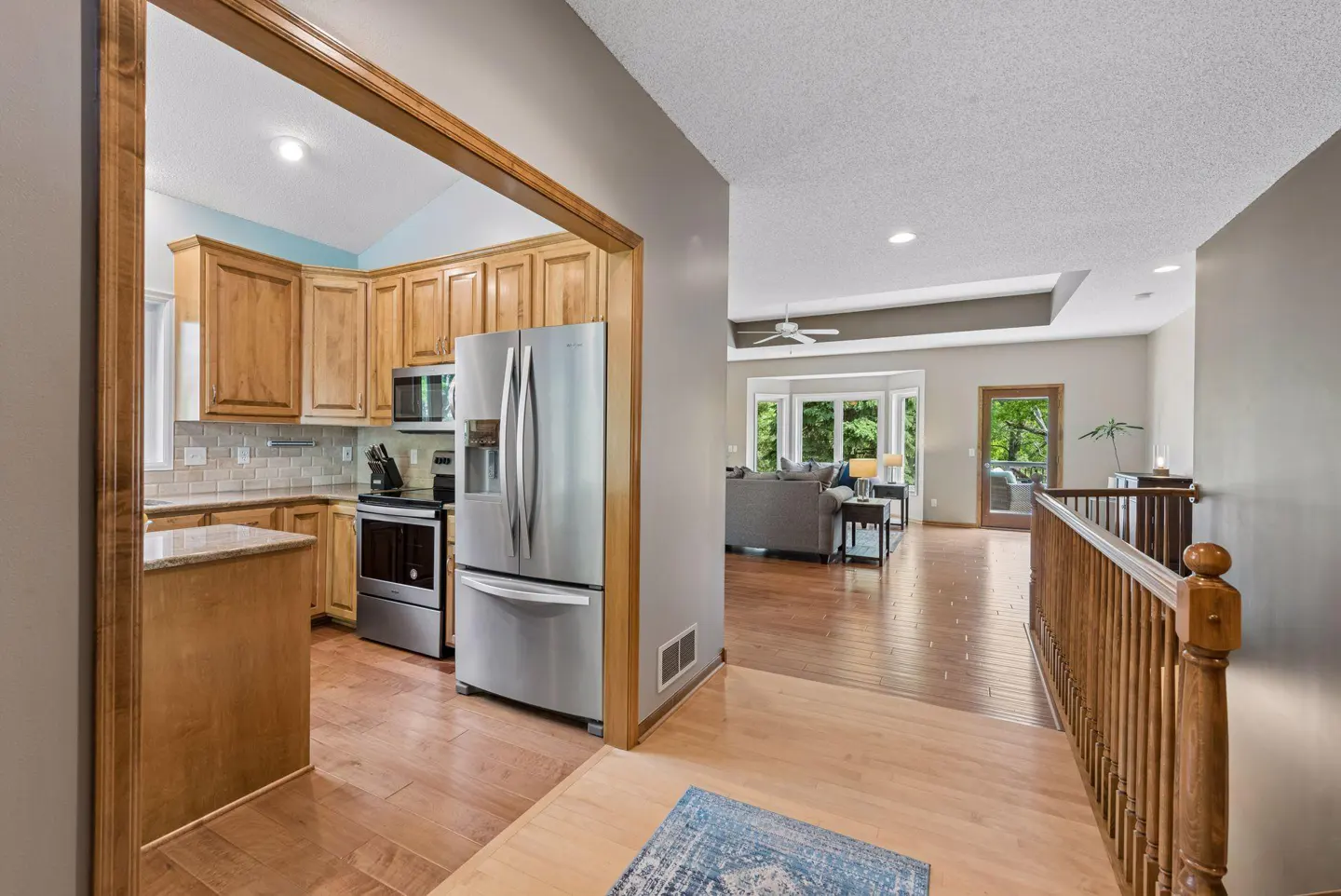 View from a hallway into a kitchen with wood cabinets and stainless appliances, opening to a living room with wood floors.