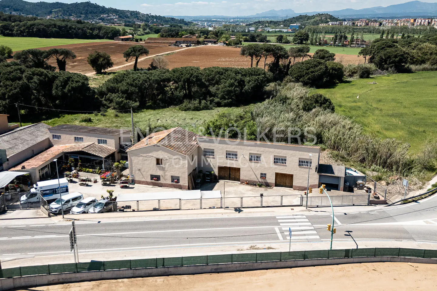 Aerial view of a commercial property with beige buildings, a parking lot with cars, and a road with a crosswalk. Fields and trees are in the background.
