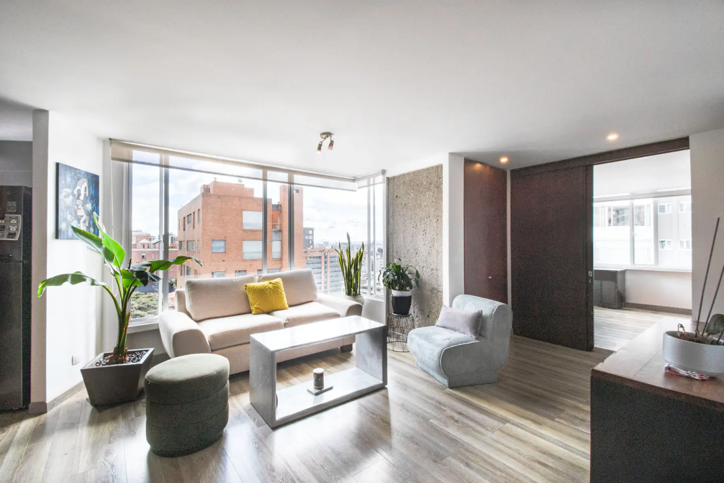 Bright living room with a beige sofa, yellow pillow, gray chair, and a large window overlooking a city.