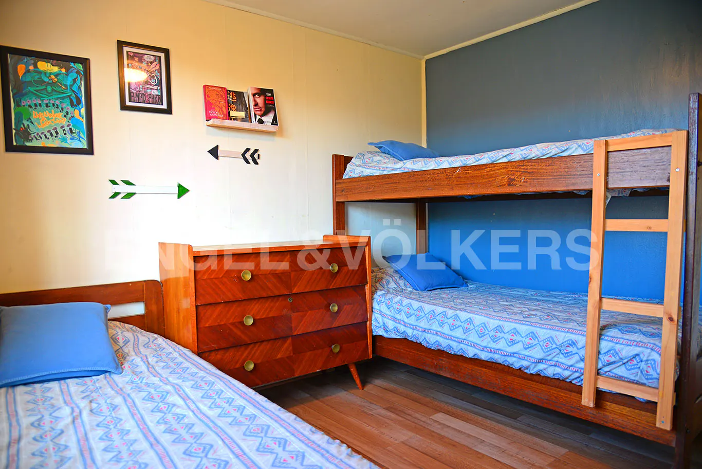 Bedroom with a wooden bunk bed, a single bed, and a wooden dresser. Blue pillows and patterned bedding. Wall art and decor.