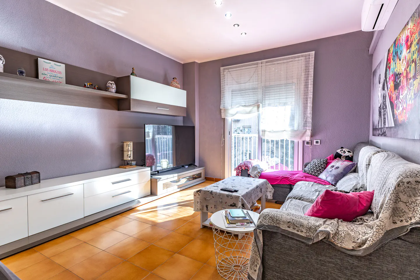 Living room with gray walls, brown tile floor, white cabinets, and a gray sofa with colorful pillows. A TV and balcony are visible.