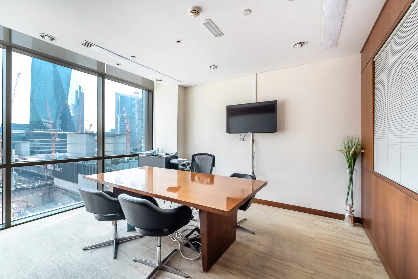 A modern office with a wood table, black chairs, and a large window overlooking a city skyline. A TV is mounted on the wall.