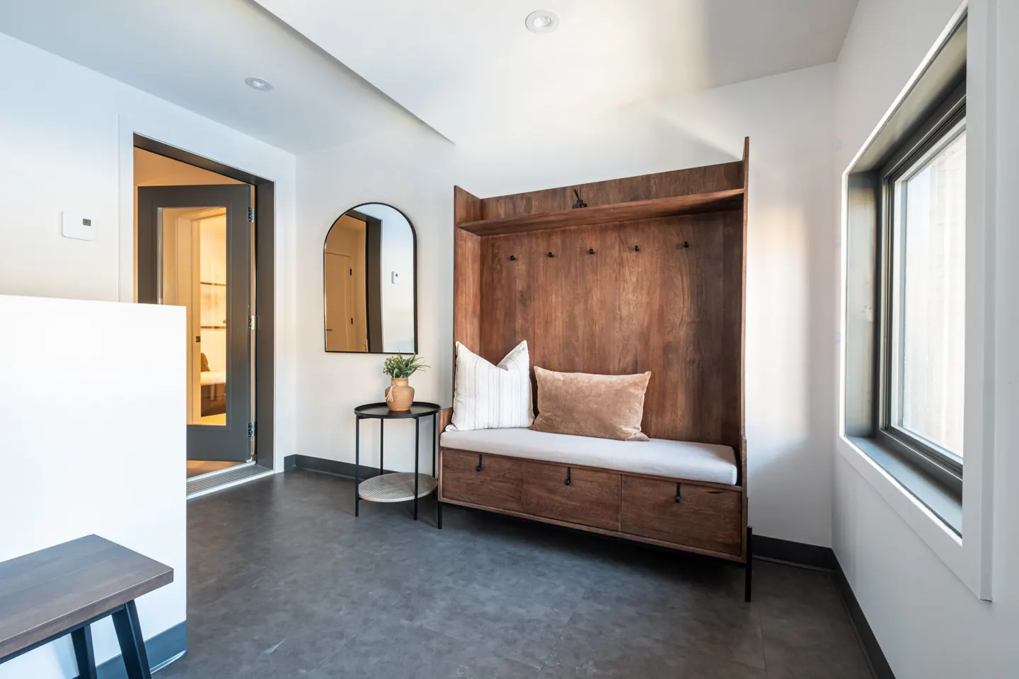 Entryway with a wooden bench and storage unit, a round mirror on a black table, and a view into another room through an open door.
