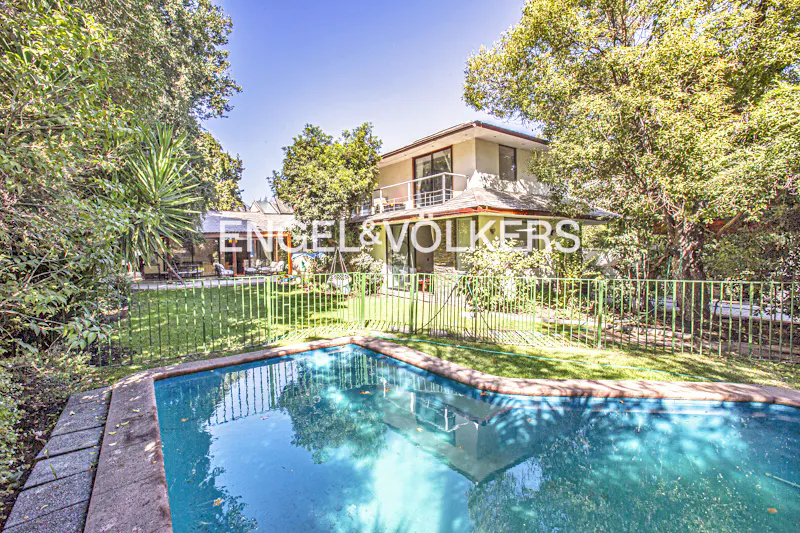 Backyard view of a two-story house with a pool, green lawn, trees, and an Engel & Volkers sign.