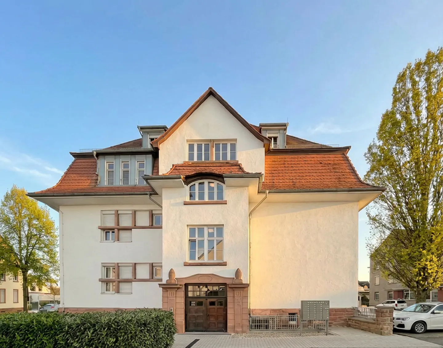 A three-story white building with a red tile roof and brown doors, surrounded by green trees and a blue sky.