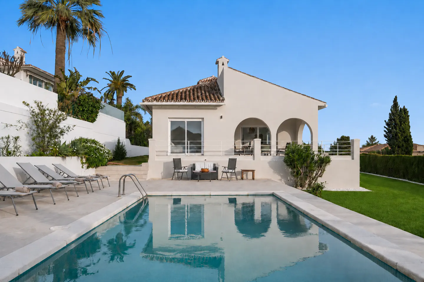 White villa with arched patio, swimming pool, and lounge chairs on a stone deck against a clear blue sky.