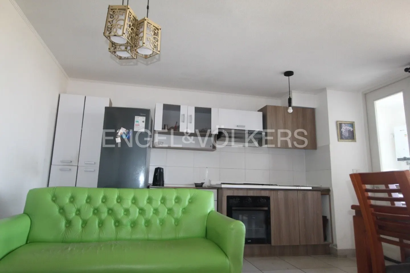 A bright kitchen with white cabinets, a stainless steel refrigerator, and a green sofa. A decorative light fixture hangs from the ceiling.