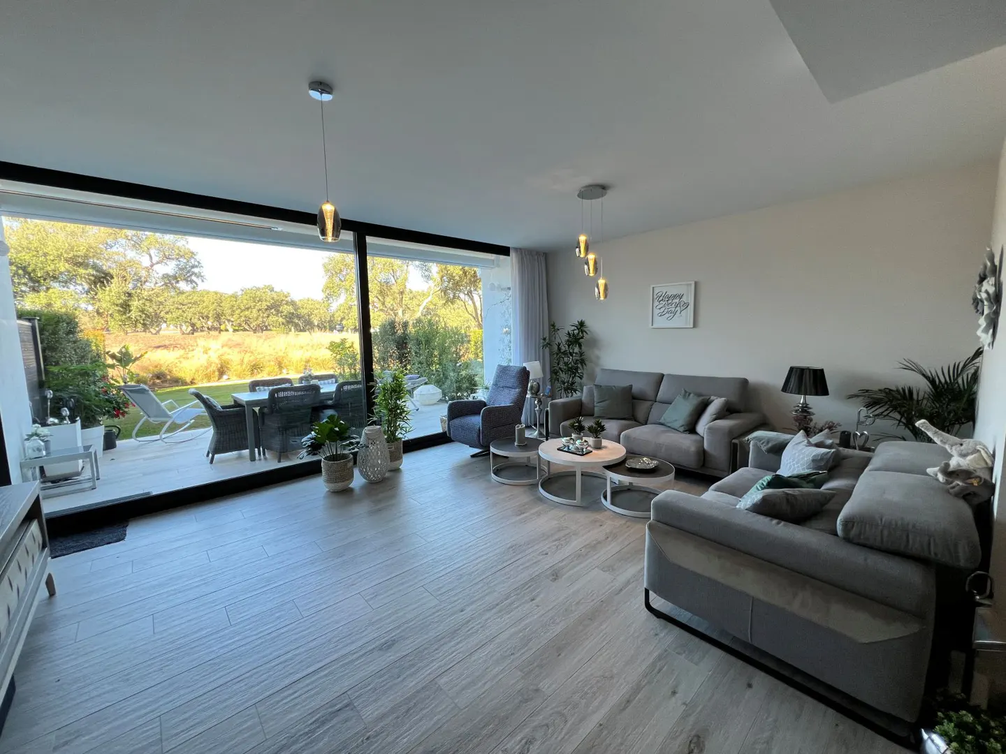 Bright living room with gray sofas, wood-look tile floor, and large windows overlooking a patio and green lawn.