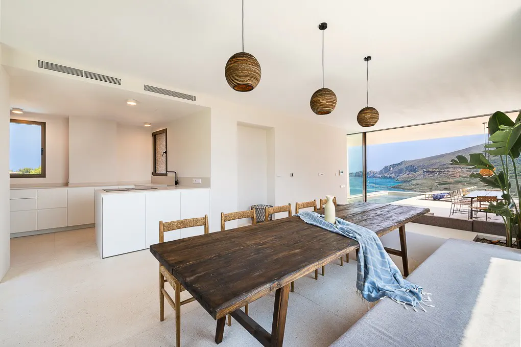 Modern dining area with a rustic wooden table, woven chairs, and pendant lights, overlooking a sea view and infinity pool.