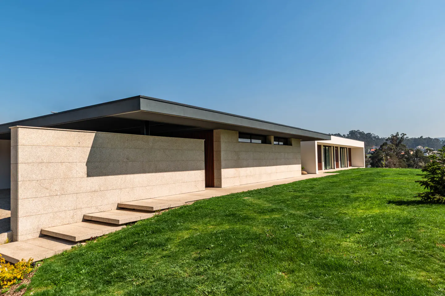 Modern, single-story home with a flat, dark gray roof, light stone walls, and a green lawn under a clear blue sky.