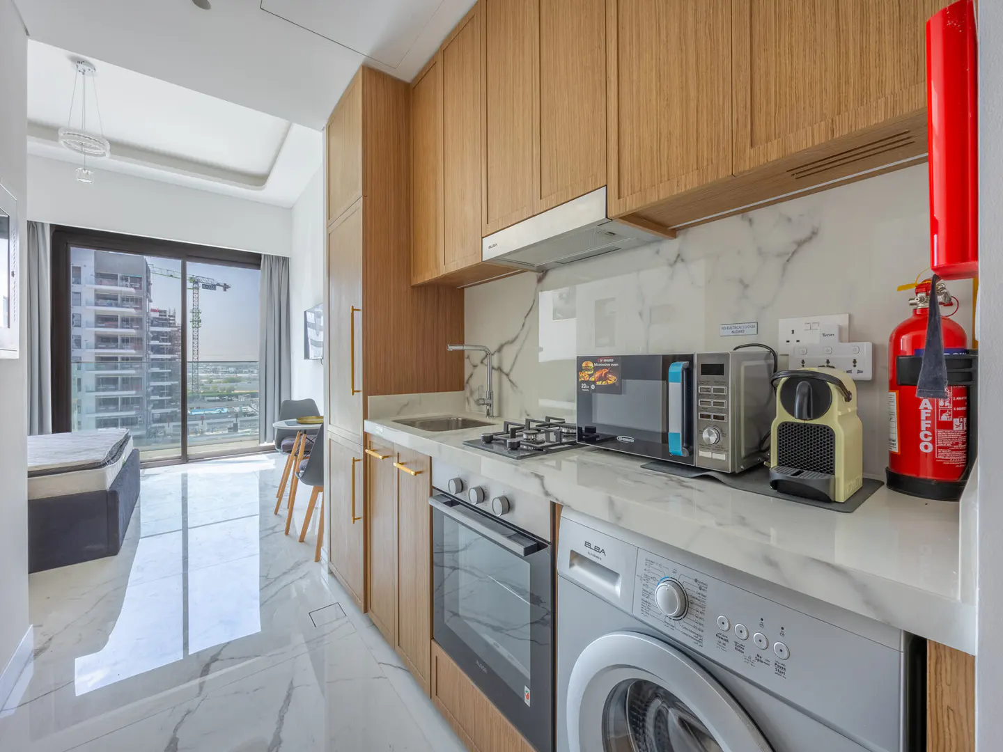 A modern kitchen with wood cabinets, marble countertops, and stainless steel appliances. A washing machine is next to the oven. A window shows a city view.