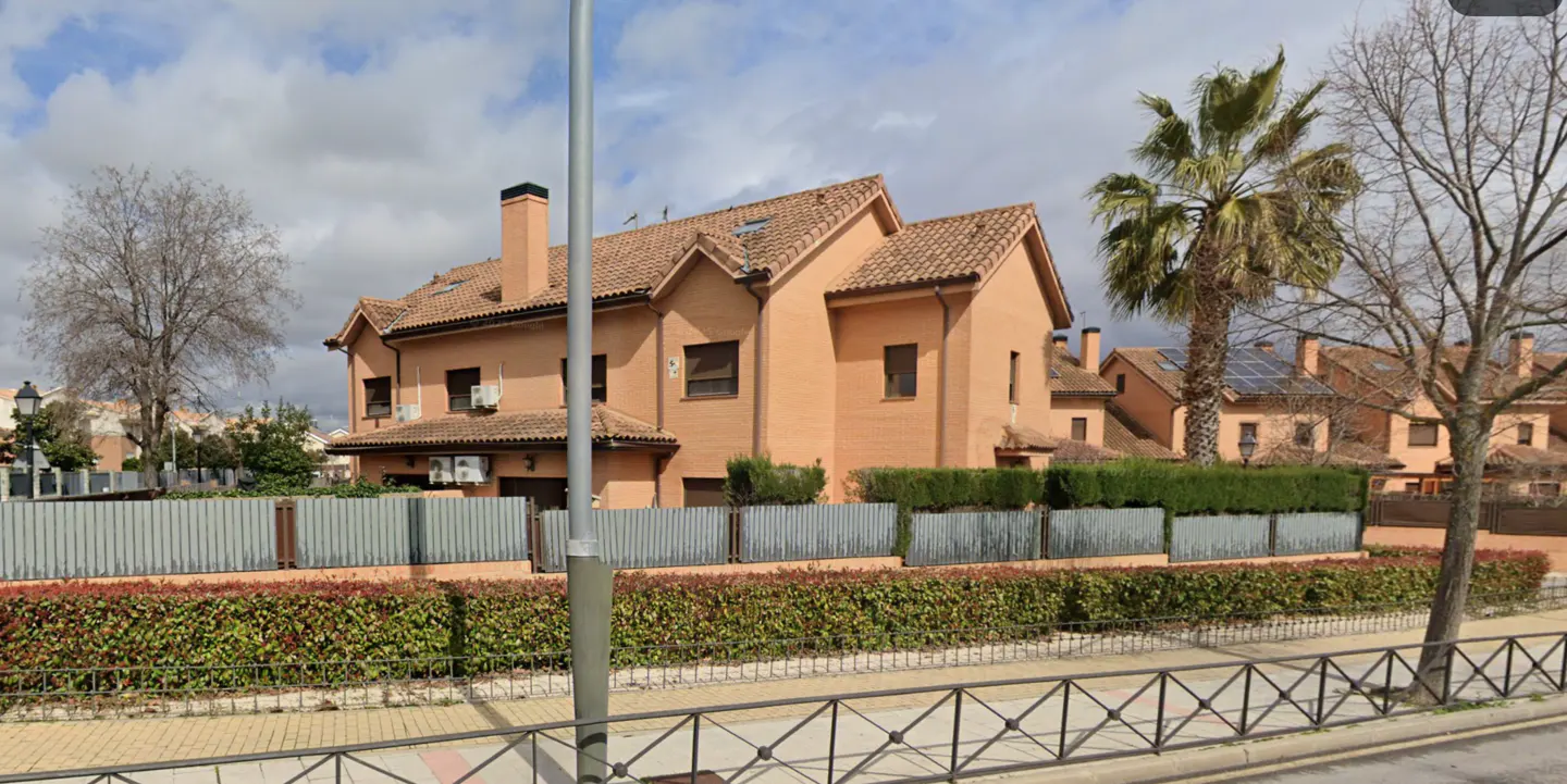 Two-story peach brick house with a brown tile roof, chimney, gray fence, and green hedge. A palm tree and bare trees are in the yard.