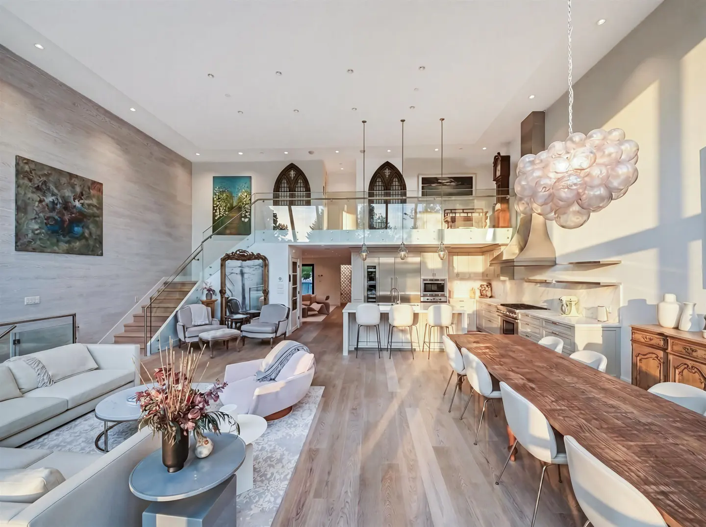 Open-concept living space with a kitchen, dining area, and lounge. Light wood floors, white walls, and modern furnishings. Artwork and a bubble chandelier add style.
