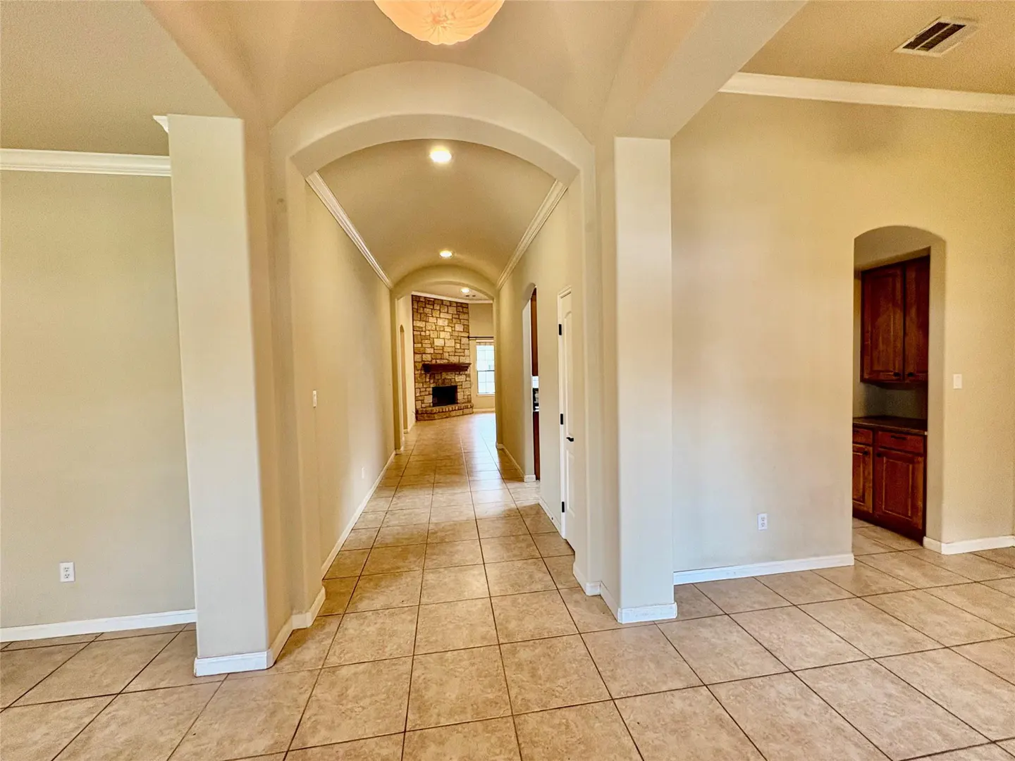 Beige hallway with arched doorways, tile flooring, and a stone fireplace visible in the distance.