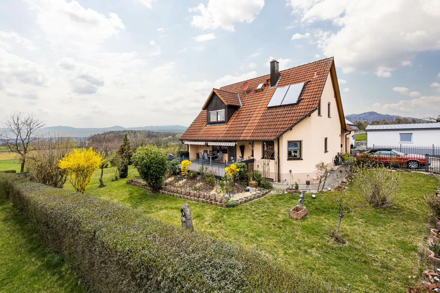 A tan house with a red tile roof and solar panels sits on a green lawn with trees and a hedge.