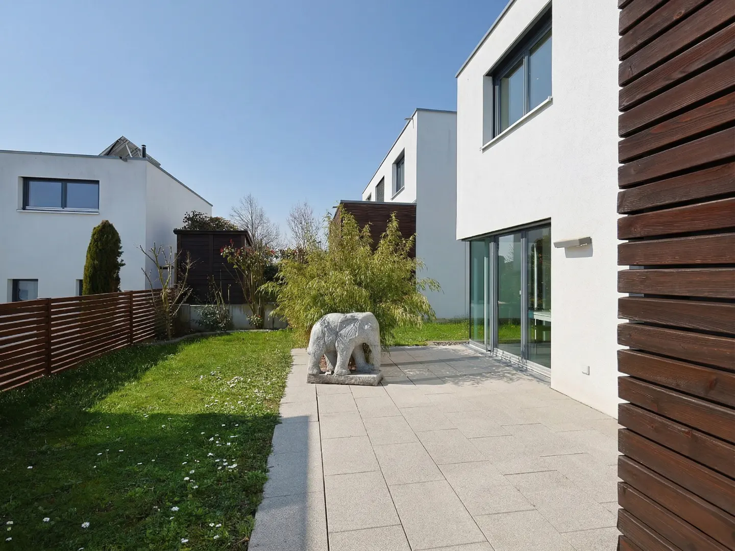 Modern white house with a stone patio, green lawn, and a stone elephant statue. A brown wooden fence is on the right.