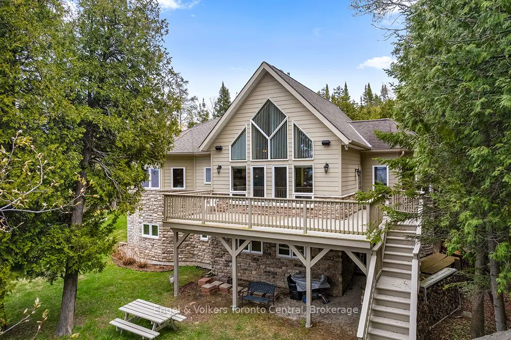 Exterior view of a beige house with a large deck, stone foundation, and tall windows, surrounded by green trees.