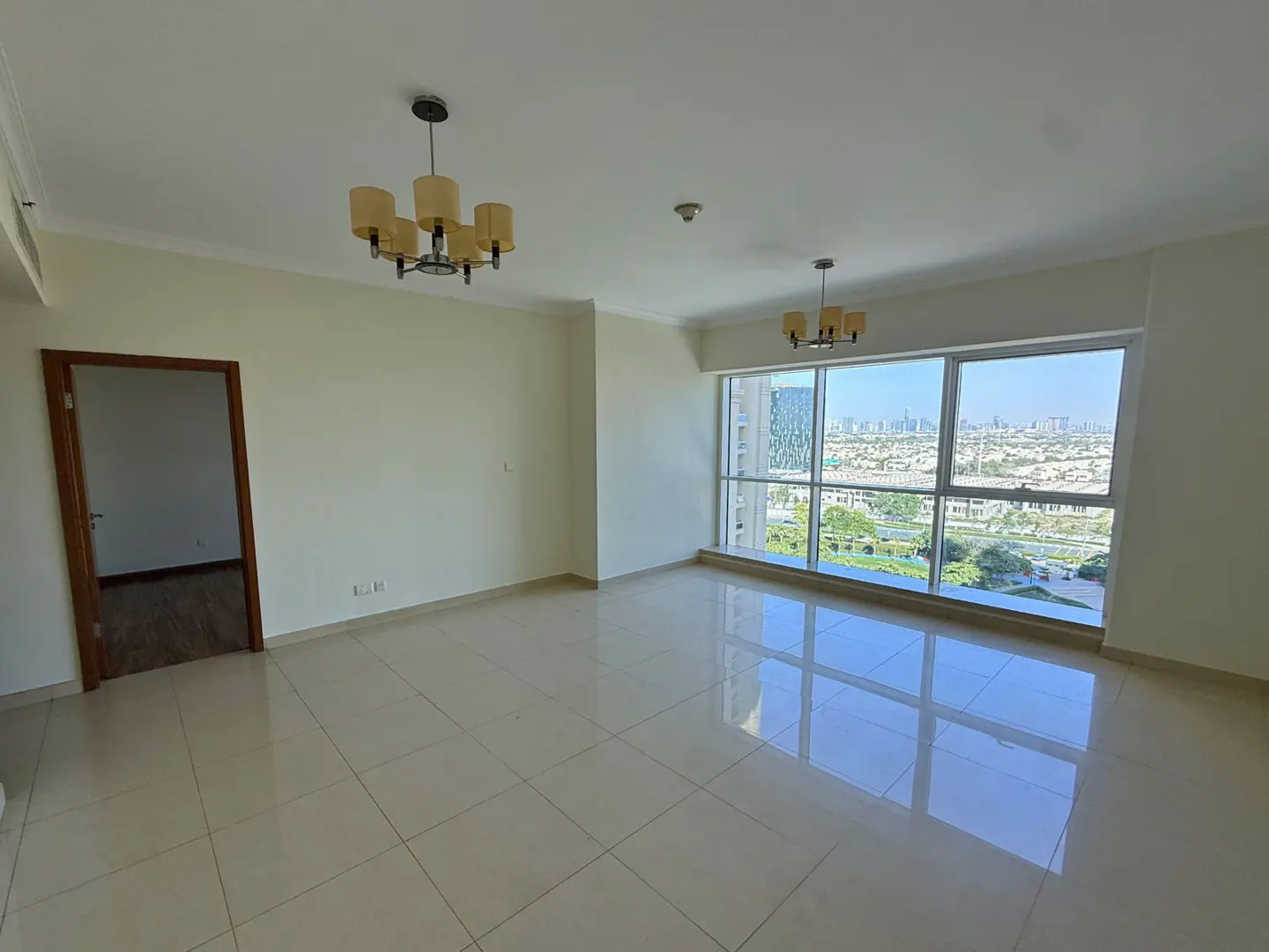 Empty, bright room with beige tile floor, white walls, and large window with city view. Two chandeliers hang from the ceiling.