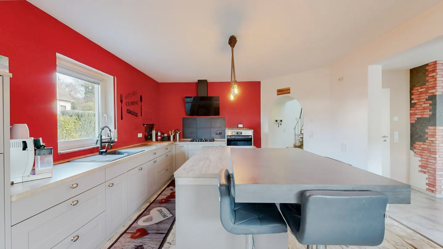 A modern kitchen with red walls, white cabinets, and a large gray island with two gray chairs. A window is above the sink.