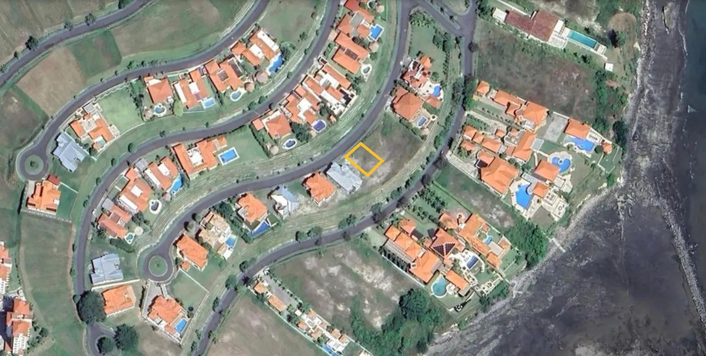 Aerial view of a residential area with large houses, pools, and a vacant lot outlined in yellow. Ocean visible on the right.