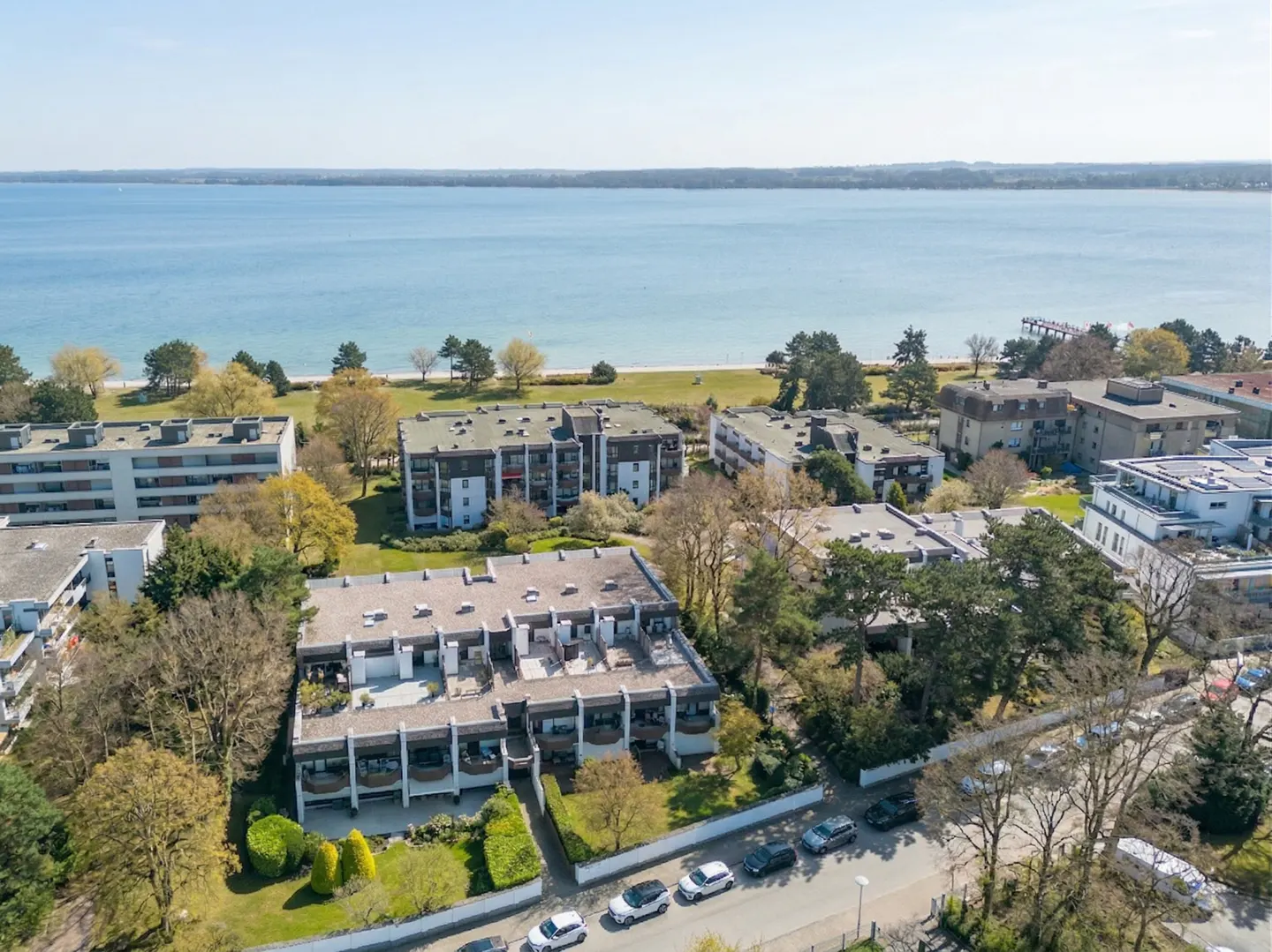 Aerial view of multi-story apartments with balconies, green lawns, trees, and parked cars near a blue ocean.