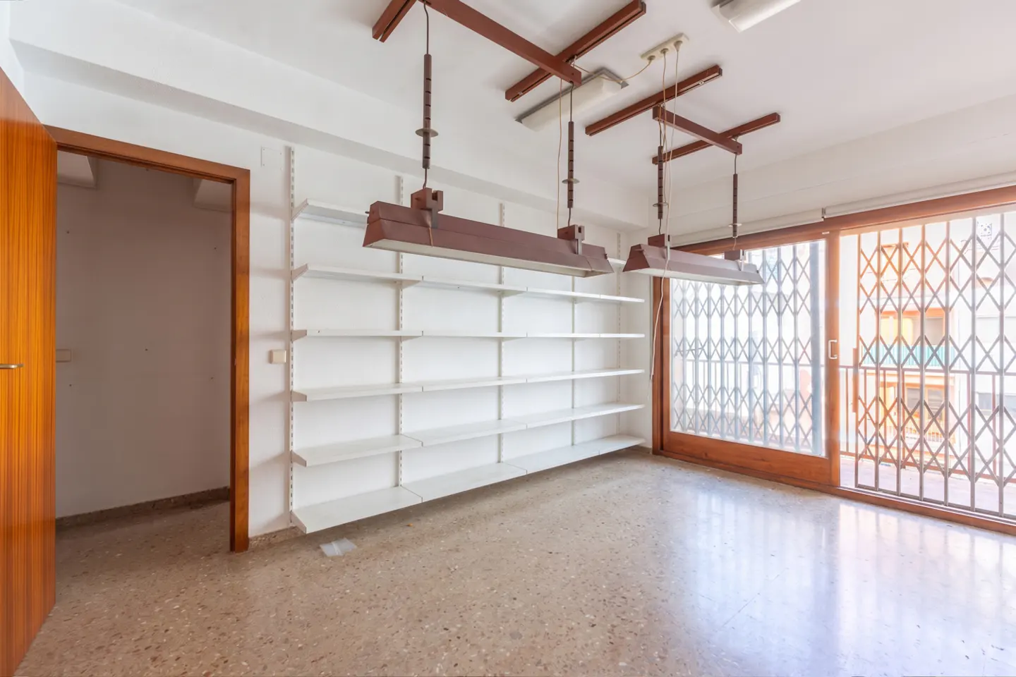 Empty retail space with white walls, terrazzo floor, and white shelving. Brown pendant lights hang from the ceiling. A wooden door and sliding glass doors are visible.