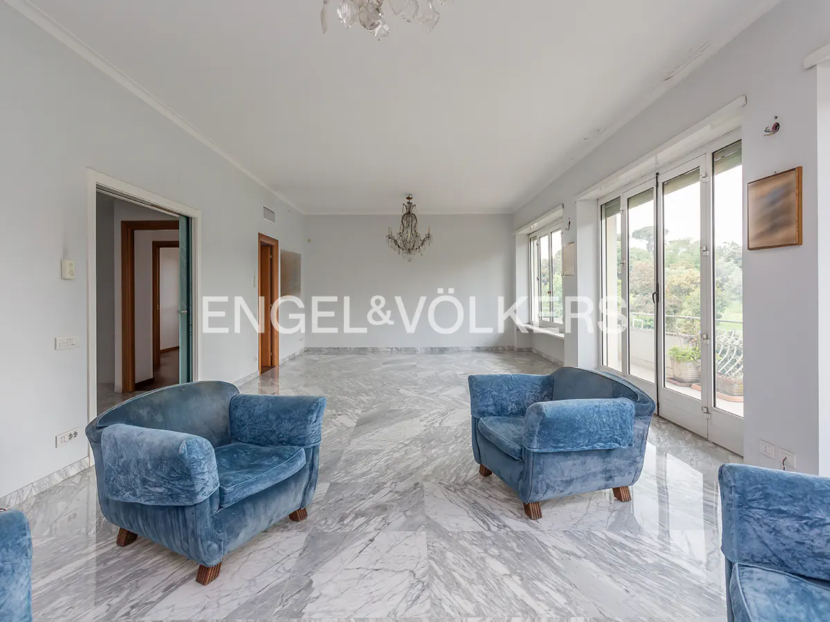 A spacious living room with blue armchairs, marble floors, and a chandelier. Windows offer a view of greenery.