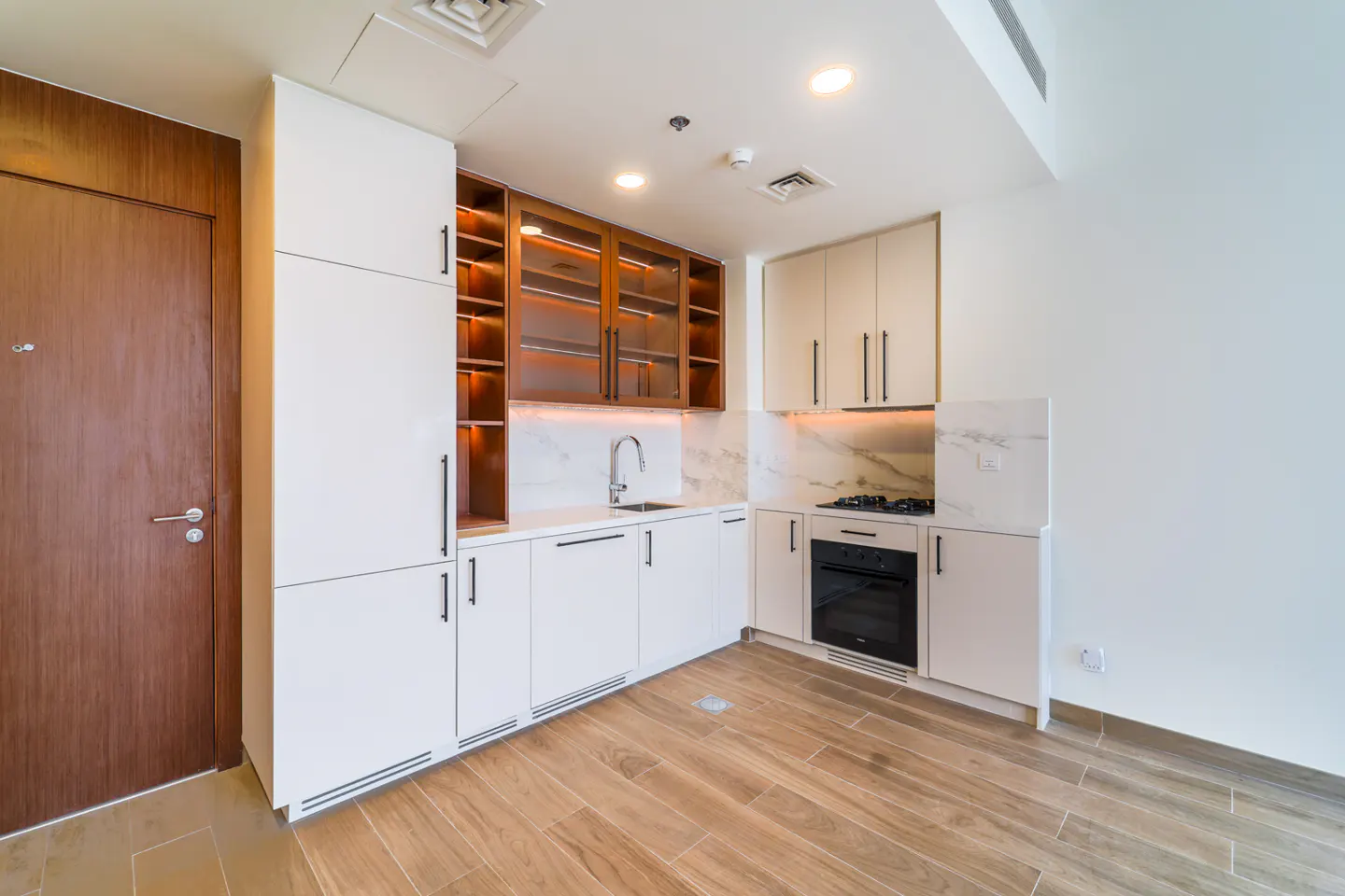 A modern kitchen with white cabinets, marble backsplash, and wood floors. A brown door is on the left.