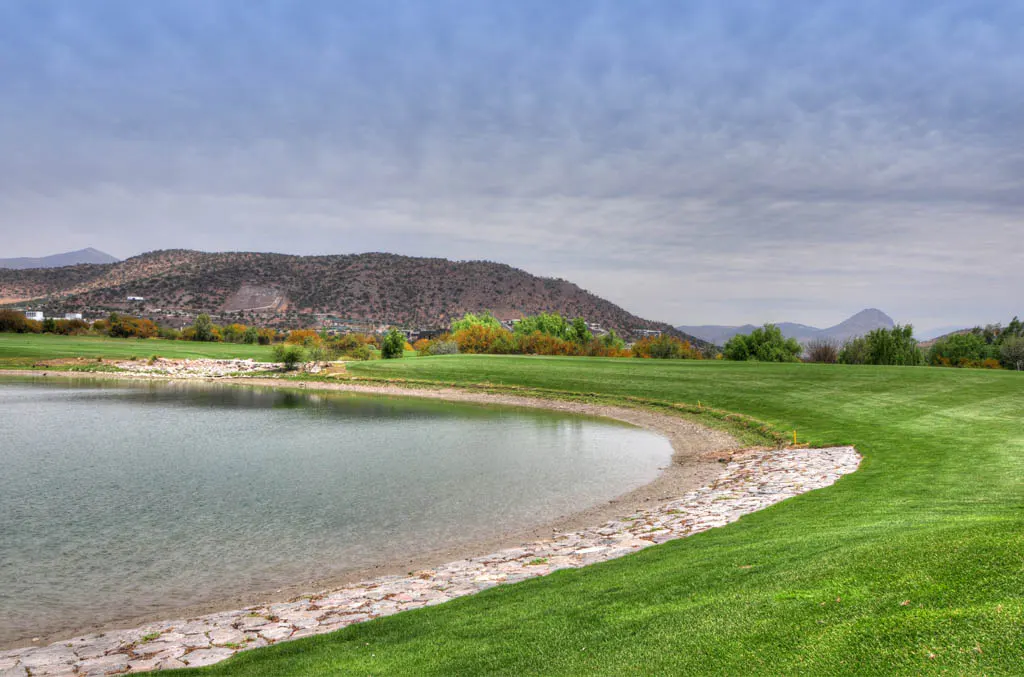A scenic view of a golf course with a pond, green grass, and mountains under a cloudy sky.