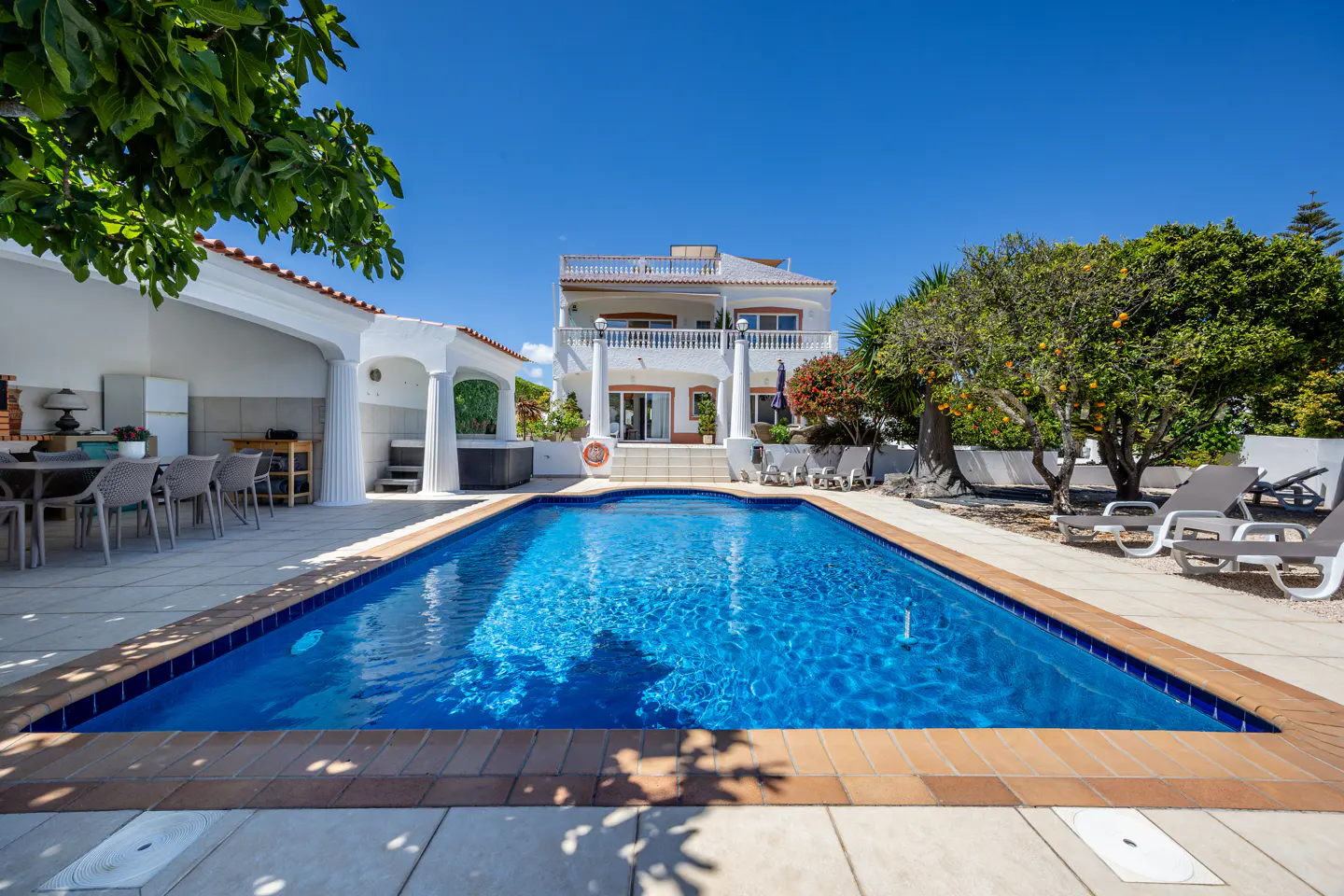 A wide shot of a two-story white house with a blue pool, patio, and lounge chairs under a clear blue sky.