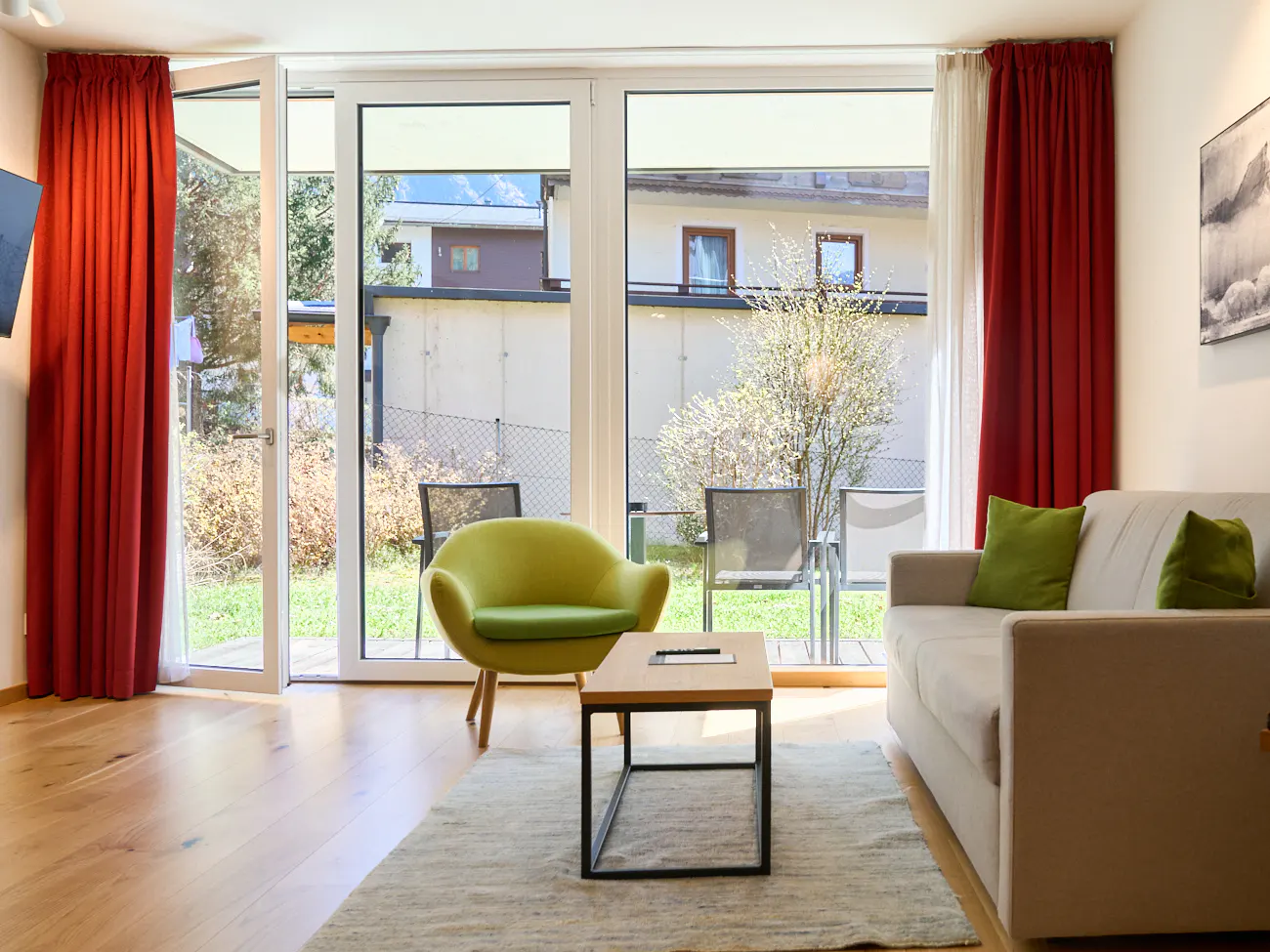 Living room with wood floors, a beige sofa with green pillows, and a chartreuse chair. Red curtains frame a glass door to a patio.