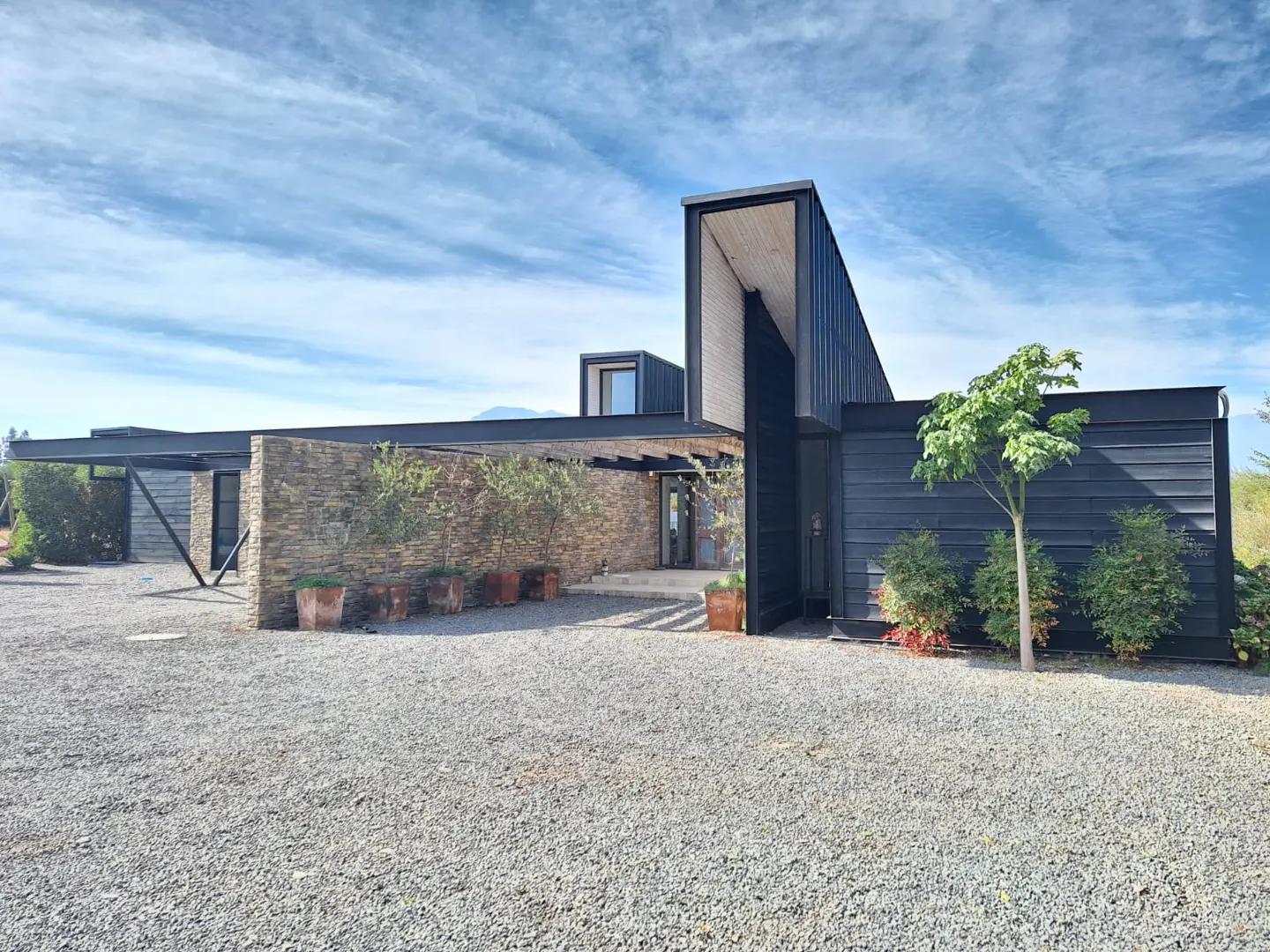 Modern home with a stone wall, black siding, and a gravel driveway under a blue sky. Trees in pots line the walkway.