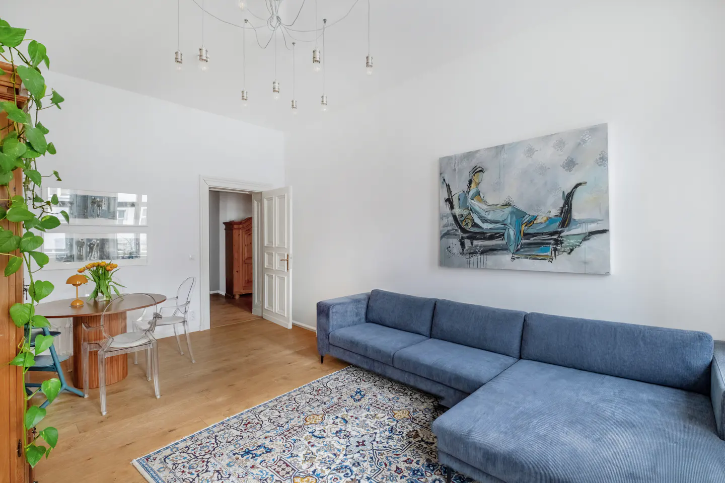 Bright living room with a blue sectional sofa, patterned rug, and a painting of a woman on a gondola. A round table with flowers sits near a vine-covered cabinet.