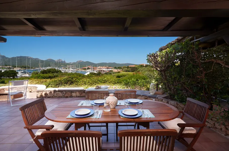 Outdoor dining area with a wooden table set for four, overlooking a marina with sailboats and mountains in the background.
