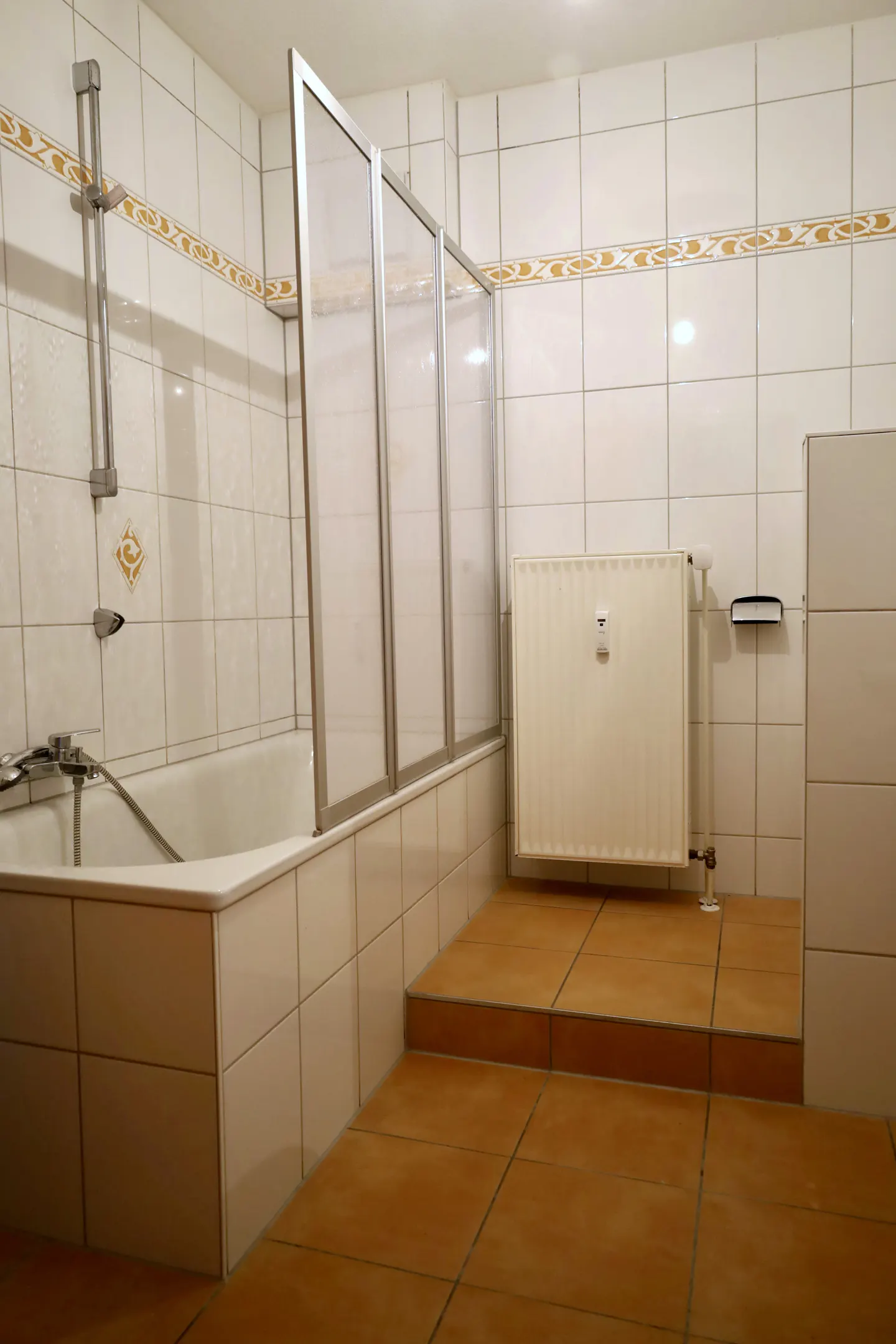 Bathroom with white tile walls, brown tile floor, bathtub with glass screen, and white radiator.