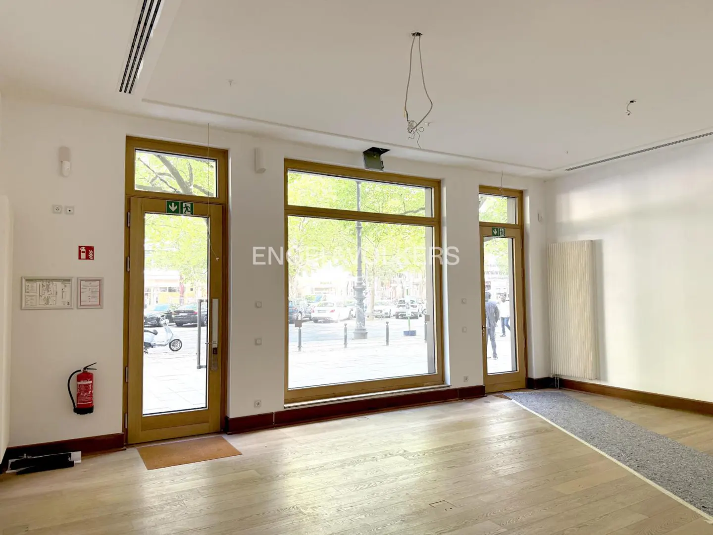 Empty retail space with light wood floors, white walls, and large windows showing a street scene with cars and trees. A red fire extinguisher is mounted on the wall.