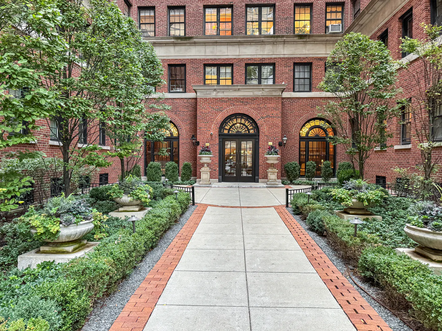 Brick apartment building with arched windows and a central entrance. A walkway leads through manicured gardens to the black double doors.