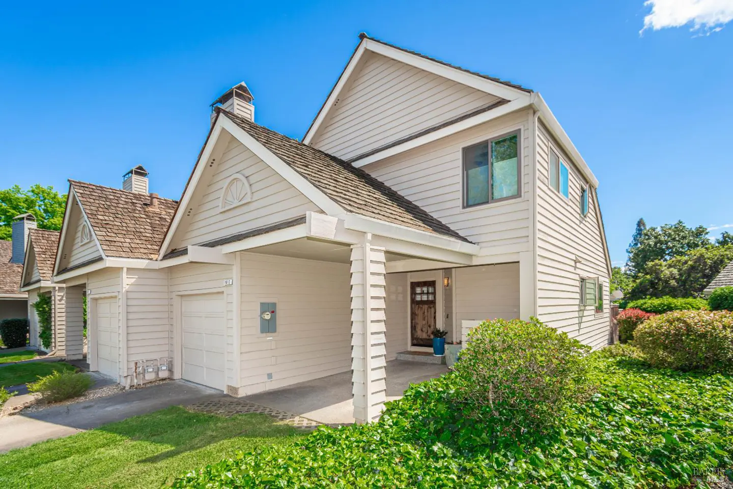 Two-story beige townhouse with a brown shingle roof, white garage door, and a carport. Green bushes in front of the house. Blue sky.