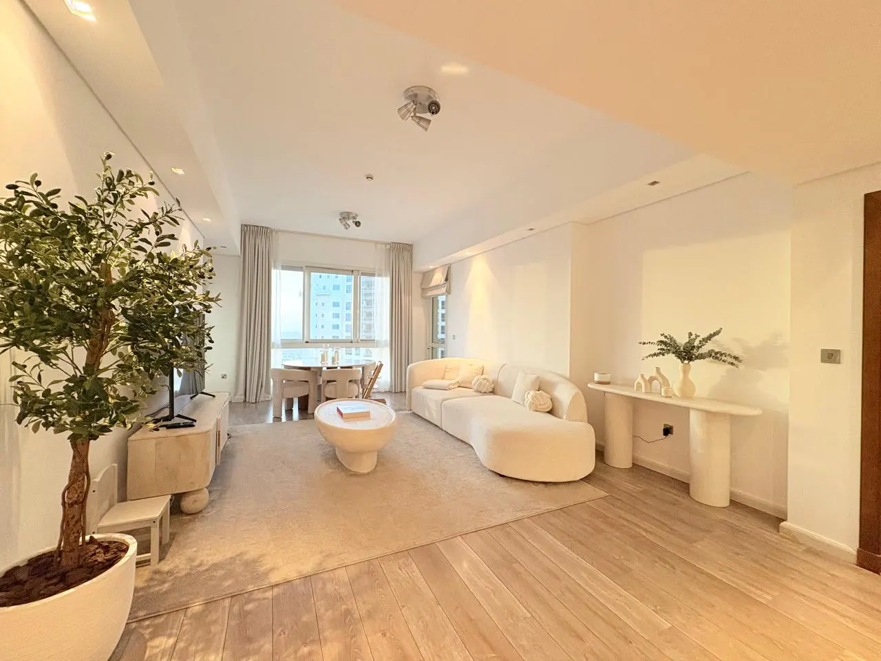 Bright, modern living room with a curved white sofa, round coffee table, and light wood floors. A large potted tree sits near a TV stand.